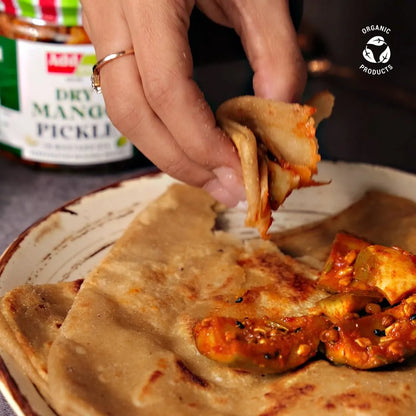 Person dipping a piece of flatbread into a bowl pickle with a jar of dry mango pickle in the background.