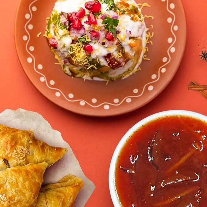 Dessert with whipped cream, pomegranate seeds, and nuts on a decorative plate, with pastries and a bowl of red sauce on an orange background.