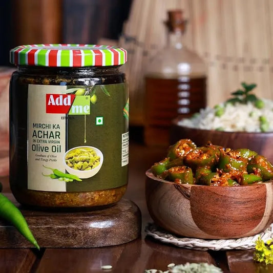 Jar of Add Me Mirchi Ka Achar with a bowl of green chilies and spices on a wooden table.